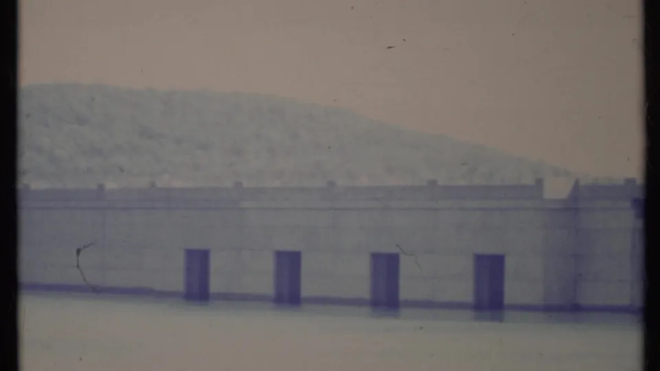 Faded 1971 California 8mm view of the dam wall across calm water, with a pale wooded slope behind it.