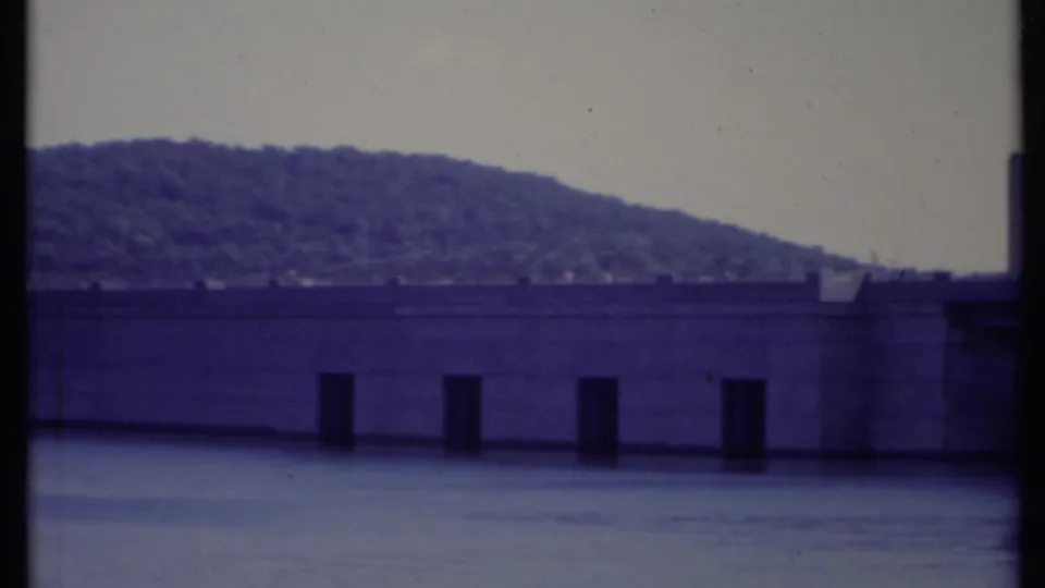 1971 California 8mm frame showing the dam face and dark spillway openings beneath a forested mountain slope.