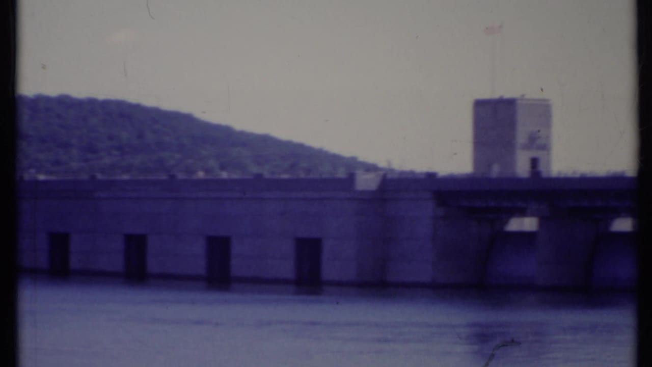 Concrete dam or bridge crossing over water with tower, hillside, and pale sky.