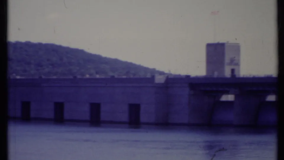 1971 California 8mm view of a concrete water-control structure across a river, with a tower and wooded hillside in the background.