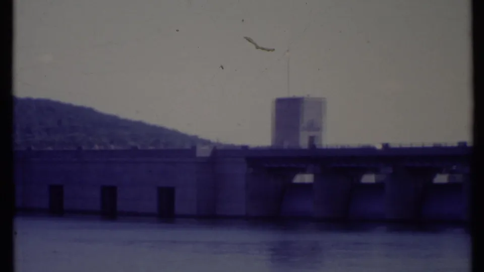 Soft 8mm frame showing a concrete dam or bridge-like water structure in California, with a tower, flag, and wooded hill beyond.