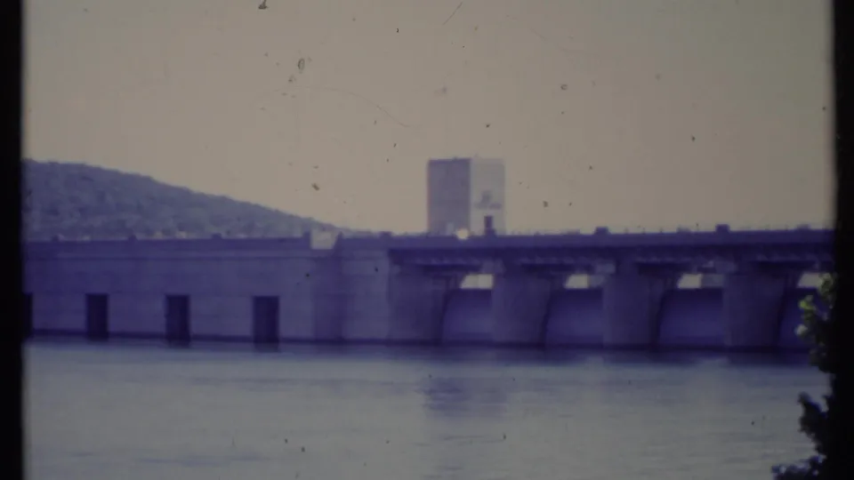 1971 8mm home-movie frame of a concrete dam or spillway across a wide river, with a tower, hillside, and foreground foliage.