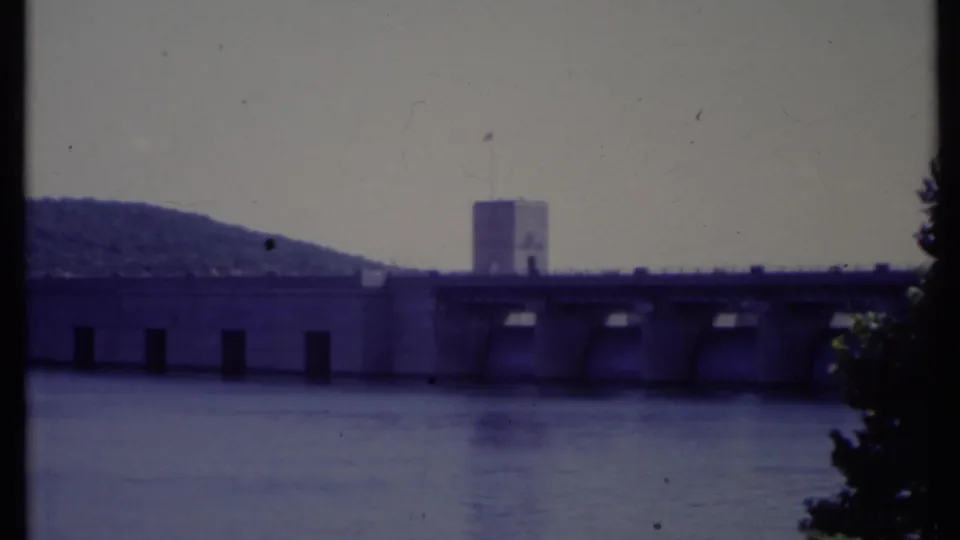 Blue-cast 8mm frame of a California concrete water-control structure, with calm river water, a tower, hillside, and dark foliage at right.