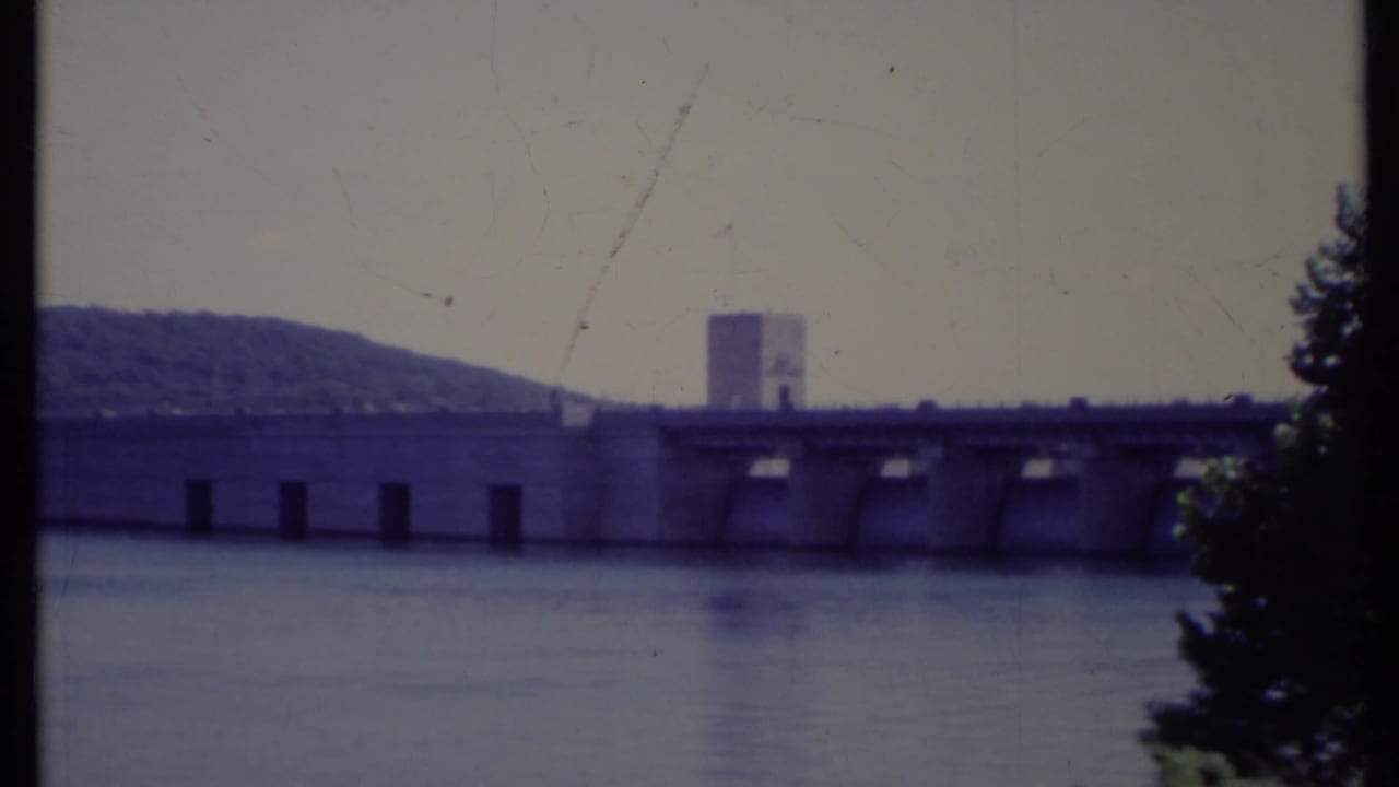 Concrete dam across wide water with wooded hill and hazy sky.