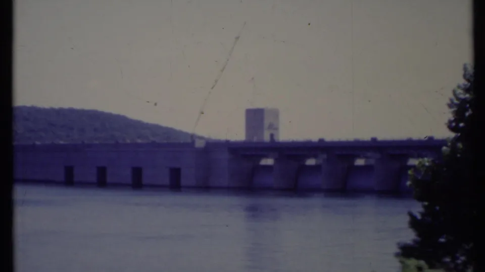 1971 California 8mm view of a concrete dam spanning wide water, backed by a forested hill under a hazy sky.