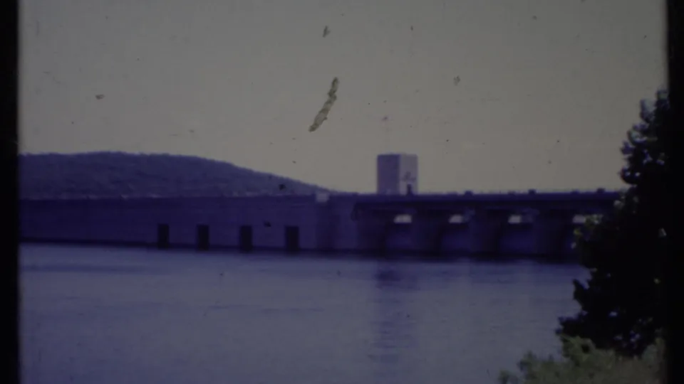 Distant 1971 California home-movie view of a dam and tower across calm water, with trees framing the right side.