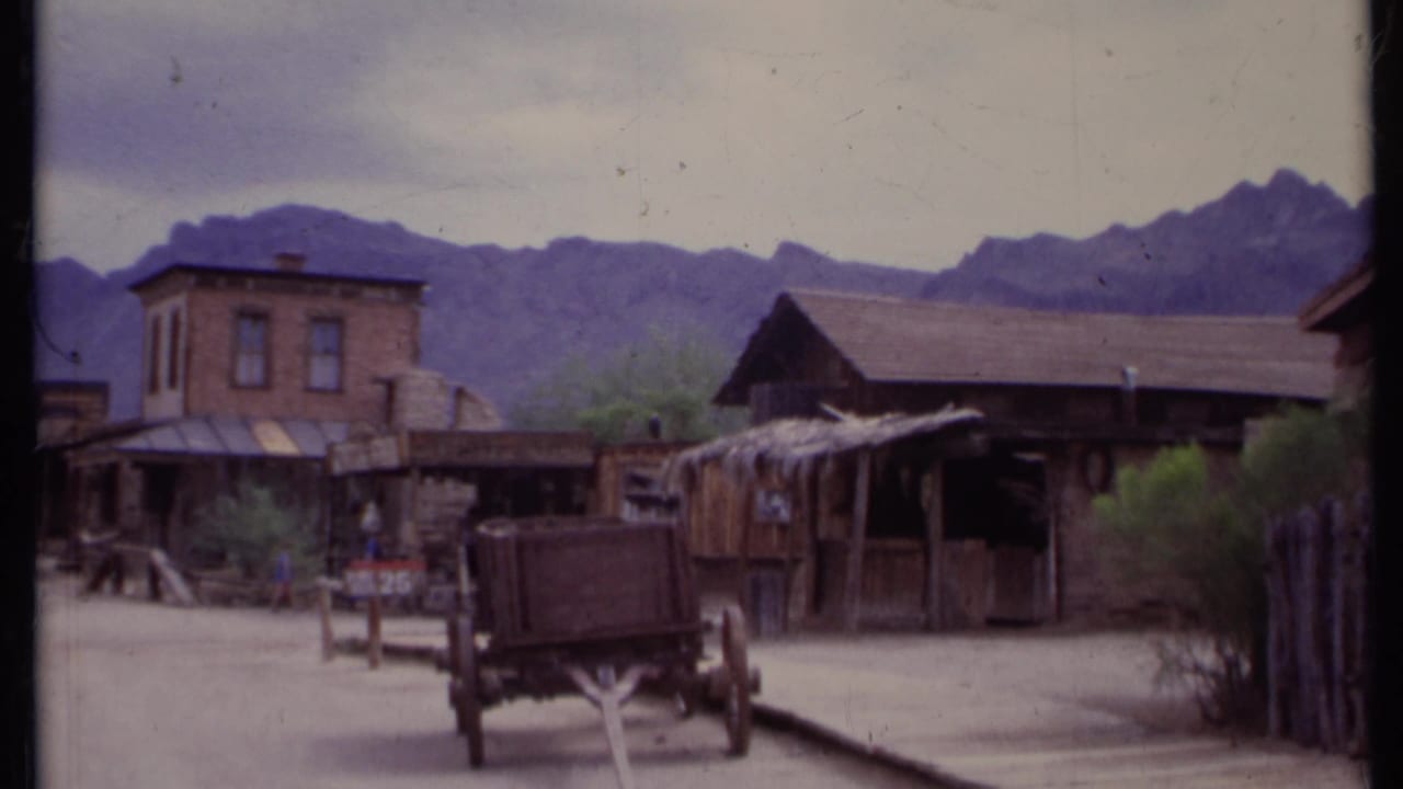 Dusty Western-style street with wooden buildings, wagon, trees, and mountains behind.