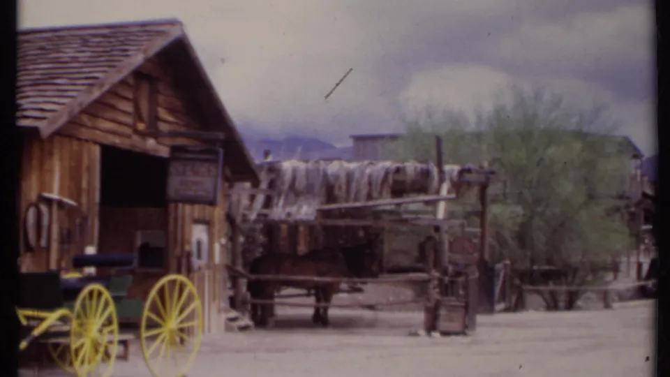 1971 California 8mm frame showing a rustic wooden stable, yellow-wheeled wagon, horse, covered wagon, and trees in an Old West setting.