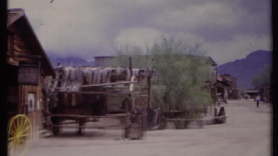 1971 California 8mm frame showing a covered wagon near a timber building, with a dusty road, trees, and mountains beyond.