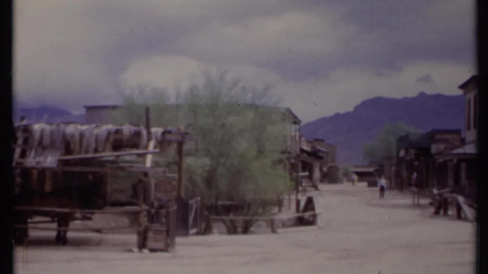 1971 California 8mm frame showing a Western-style town road with wagon, trees, storefronts, distant visitors, and mountains under clouds.