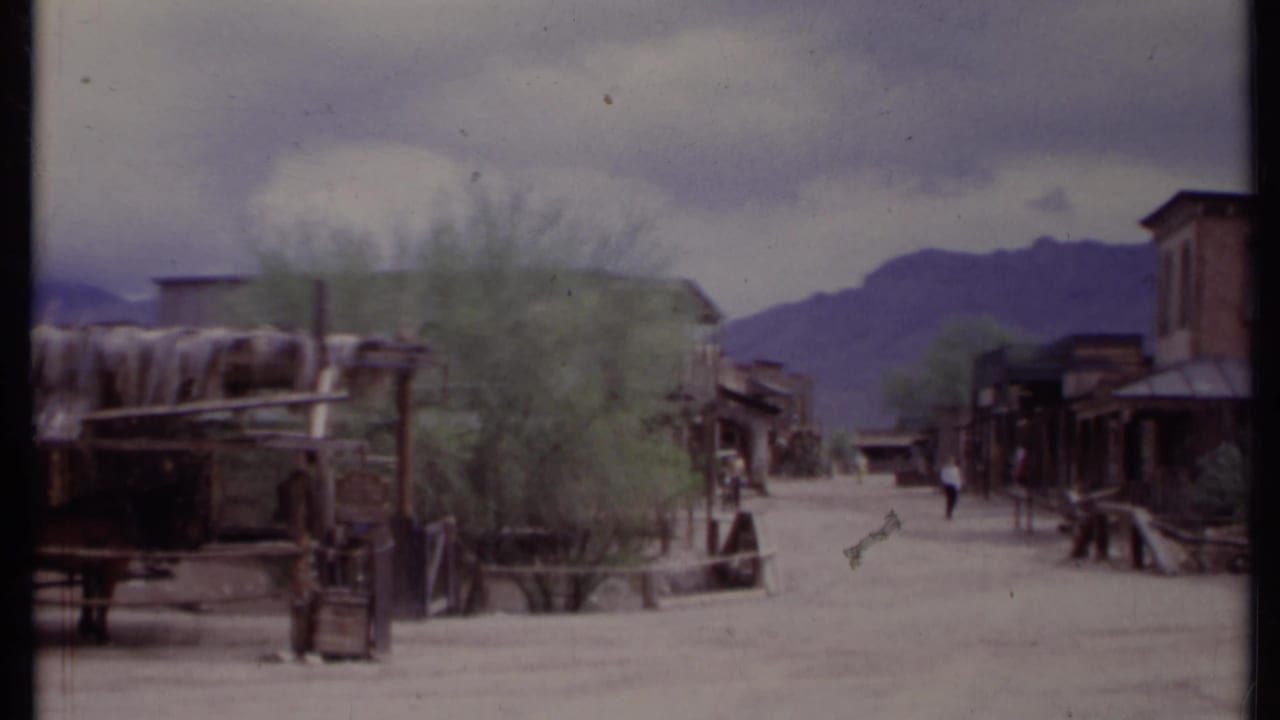 1971 California 8mm frame of a recreated Old West-style street with wooden storefronts, wagon elements, pedestrians, and mountains beyond.