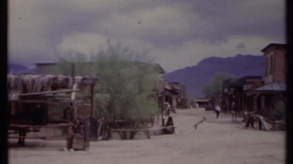 1971 California 8mm frame of a recreated Old West-style street with wooden storefronts, wagon elements, pedestrians, and mountains beyond.