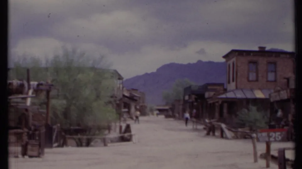 1971 California home-movie frame showing visitors on a dusty Old West-style street with wooden buildings, wagon elements, signs, and mountains.