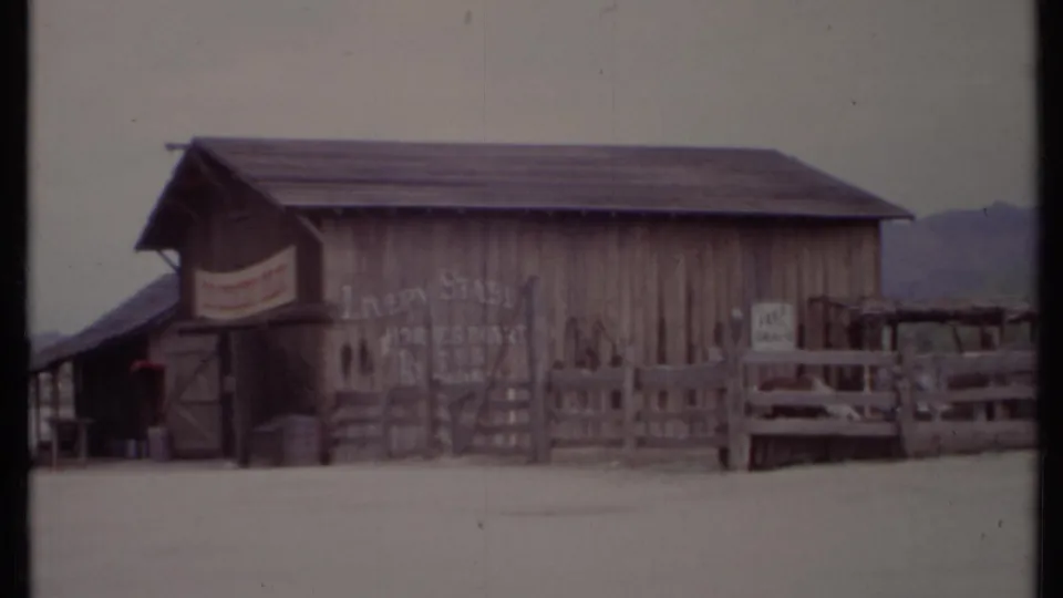 1971 California 8mm frame of a barn-like Old West-style building with wooden fencing, signs, barrels, and a mountain backdrop.