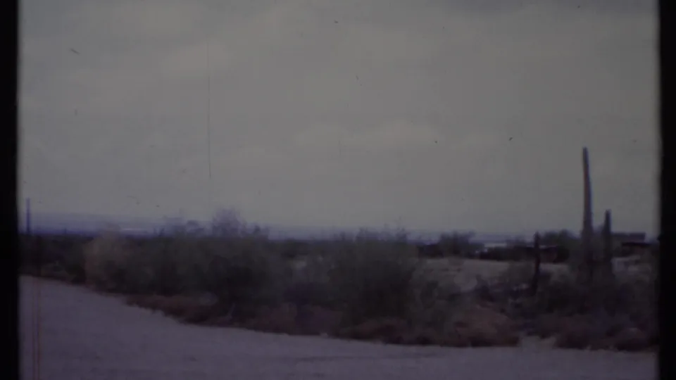 1971 California desert roadside with a pale dirt road, scrub brush, tall cacti, and a cloudy sky in soft 8mm color.