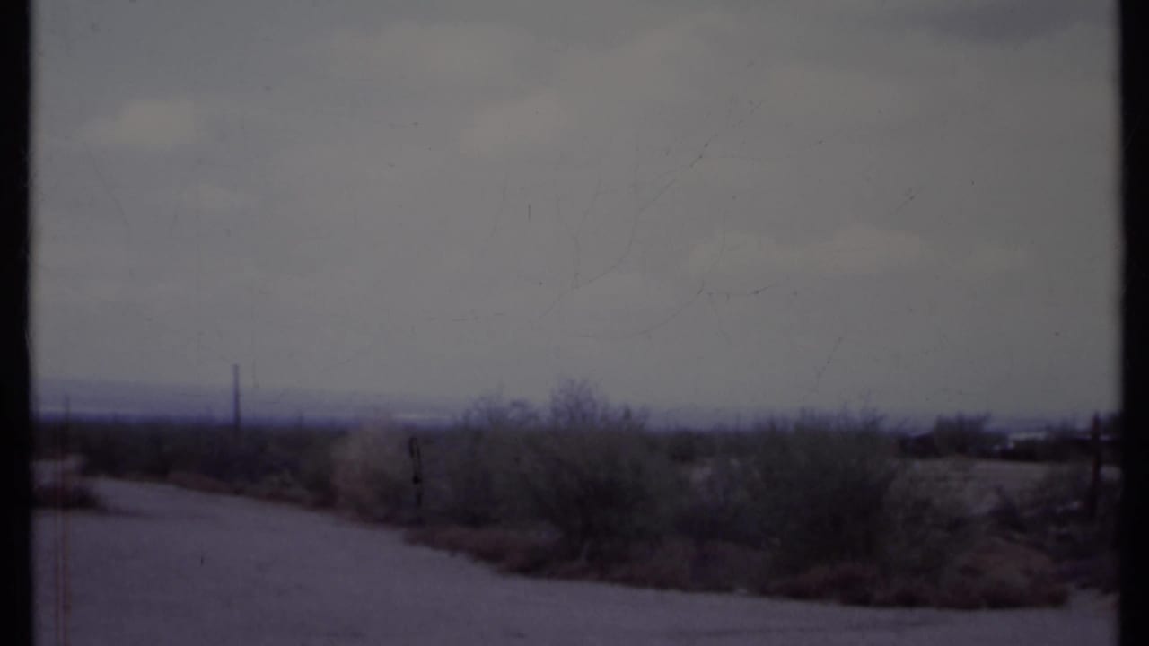 Open desert road with scrub vegetation, distant poles, and a cloudy sky.