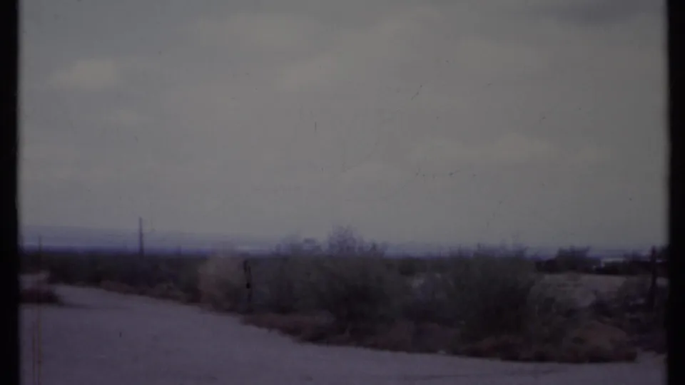Sparse California desert view from 1971 home-movie footage, showing road surface, brush, distant poles, and overcast sky.