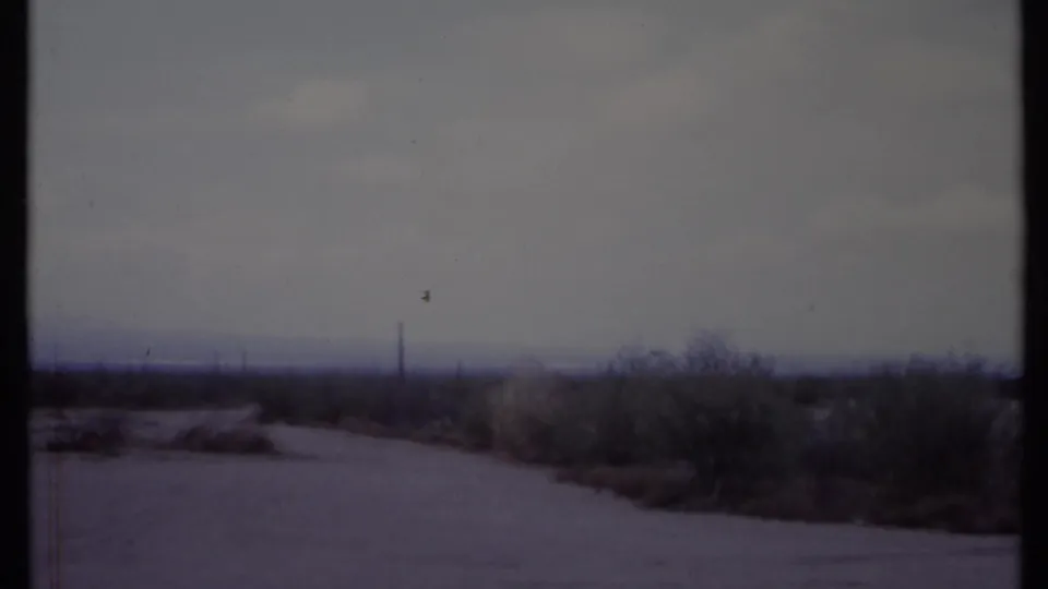 Muted 8mm frame of a California desert roadside in 1971, with pale road, low brush, distant utility pole, and heavy sky.