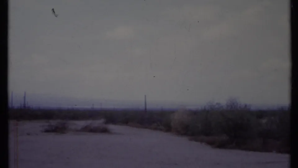 California desert road in 1971 8mm footage, bordered by scrub brush with faint poles along the horizon under a cloudy sky.