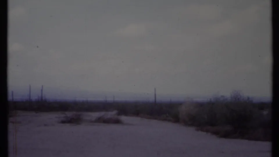 1971 California home-movie view of a pale dirt road crossing sparse desert-like scrubland with utility poles on the horizon.