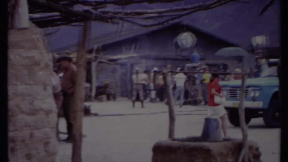 A 1971 California roadside gathering appears near a wooden structure, with several people, a blue truck, and rustic posts in view.