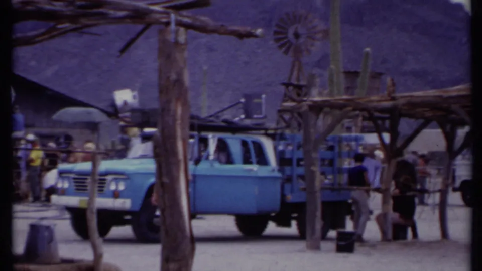 A clearer 1971 California frame of a blue truck parked by a wooden desert set while crew and bystanders stand near equipment.