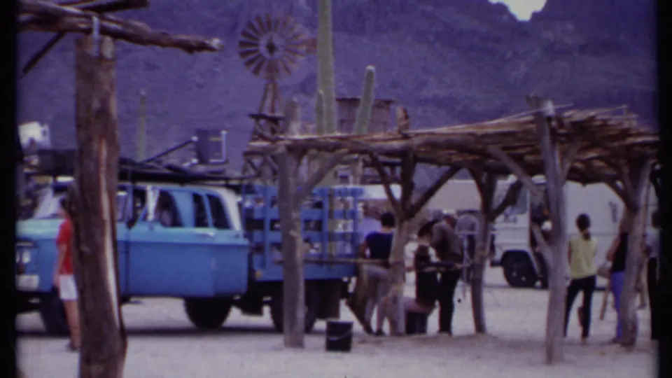 1971 California 8mm frame with bystanders and crew near a blue truck, wooden shelter frames, cacti, equipment, and mountains.