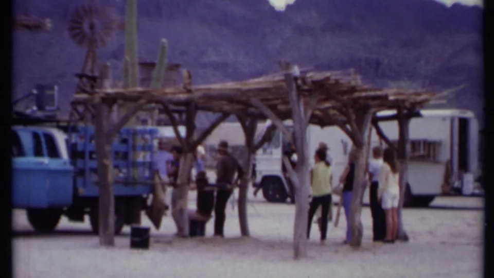 A 1971 California desert set view showing people gathered under wooden structures, with a blue truck, white vehicle, cacti, and mountains behind.