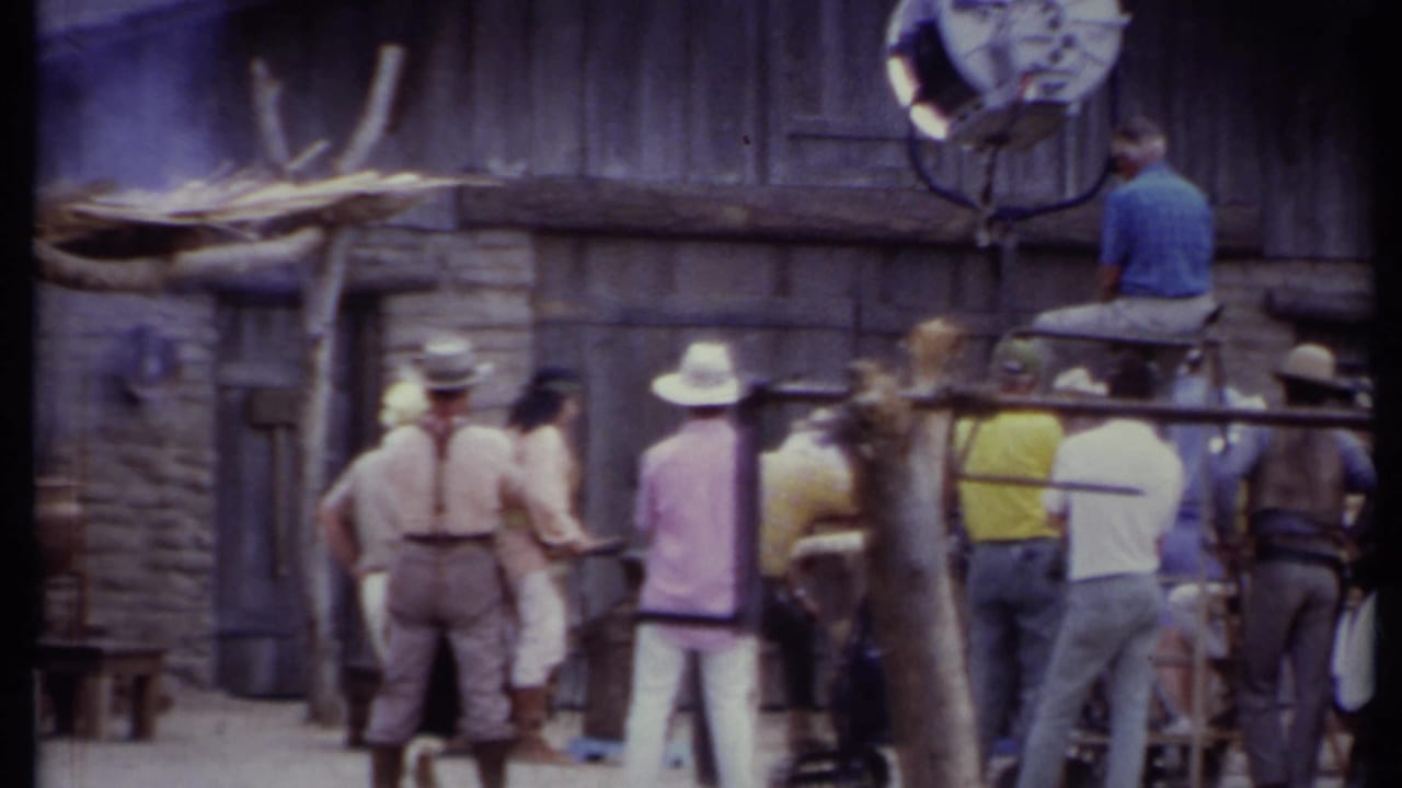Crew members gather near a rustic Western set facade with a large film light overhead.