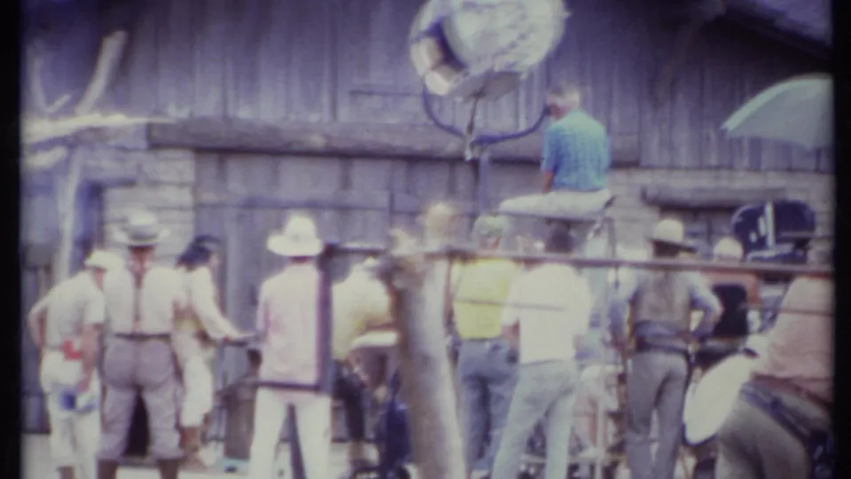 Crew members in hats and casual clothing stand around a Western-style set in California, with a large light and seated man above.