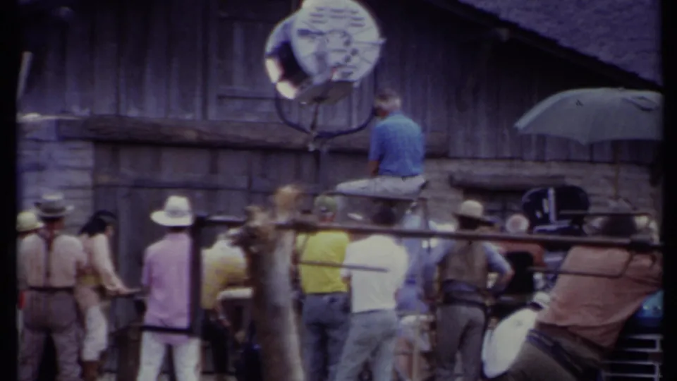 Soft 8mm frame of a Western shoot setup, with crew members, hats, a high light, and production equipment in view.