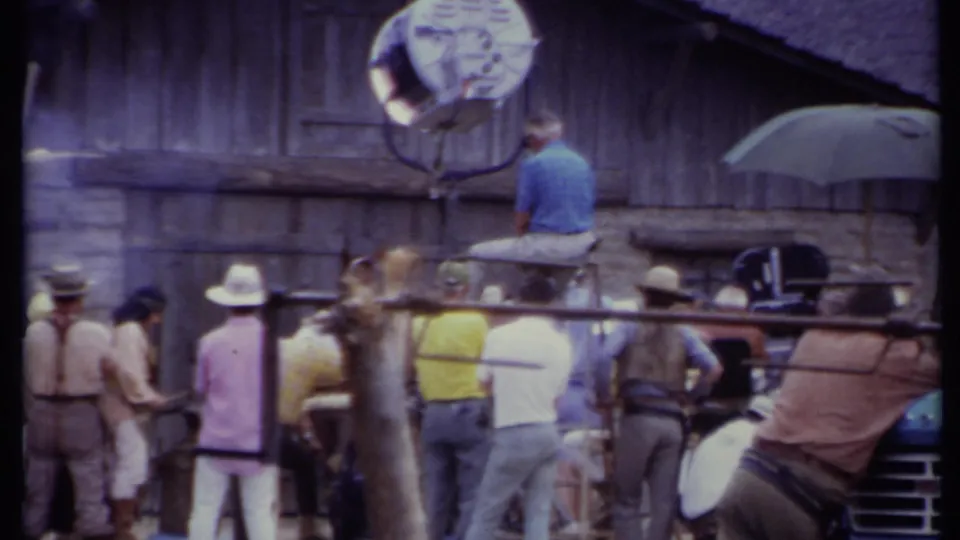 1971 California behind-the-scenes frame with a Western set facade, crew members, lighting rig, and partial vehicle visible.