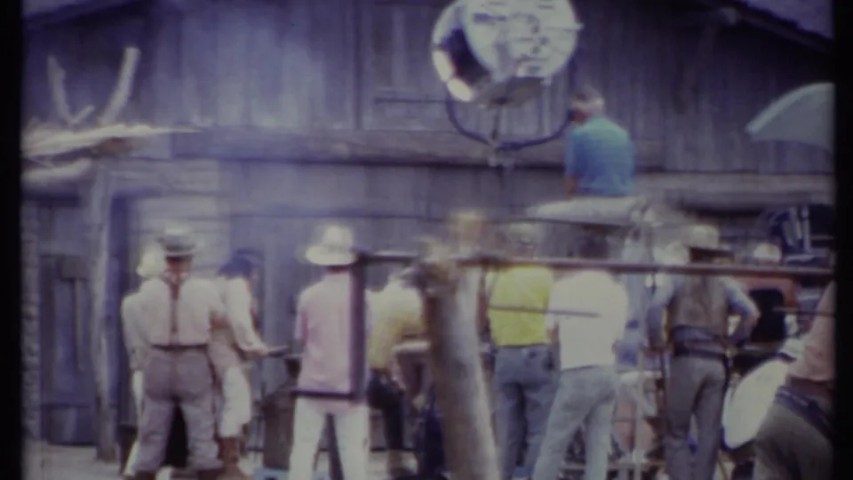 1971 California 8mm frame with people in hats and casual clothing near a Western barn set and visible production lamps.