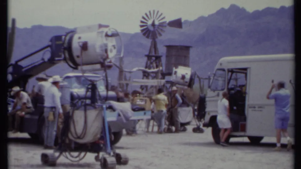 1971 California 8mm frame showing a film light, windmill prop, vehicles, crew, and mountains behind a Western-style set.