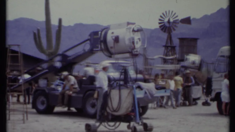 1971 California 8mm frame with a large studio lamp, windmill, cactus form, vehicles, and crew against desert mountains.