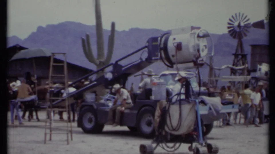 1971 California 8mm frame showing crew and large production lights on a Western-style desert set with cactus, windmill, and mountains.