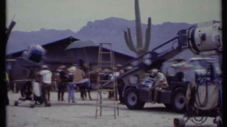 1971 California frame with crew, horses, camera or lighting gear, a cactus, and wooden set structures in a desert Western production setting.