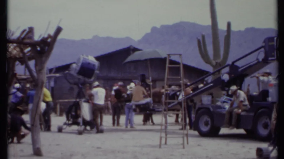 1971 California 8mm frame showing a busy Western-style set with crew, large lights, ladders, horses, cactus, and desert mountains.