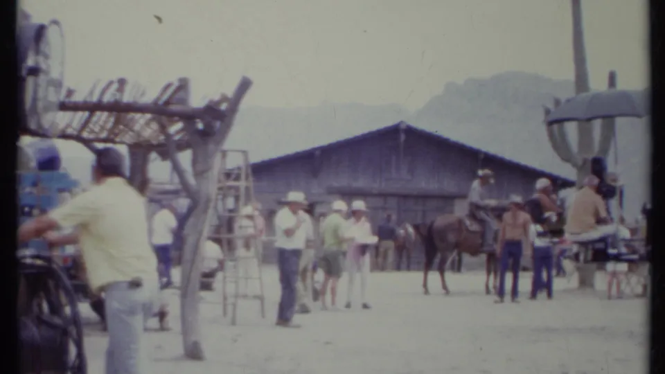 1971 California 8mm frame showing crew and horses near a rustic Western set building, with ladders, cactus, and pale desert mountains behind.