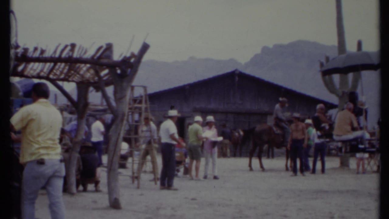 Outdoor Western film set with people, horse, ladders, wooden structures, and mountains behind.