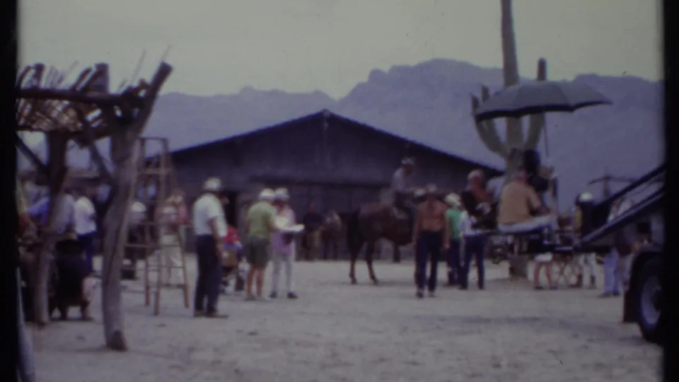 1971 California frame with a rider on horseback beside crew members, a sun umbrella, ladders, set buildings, and mountains.