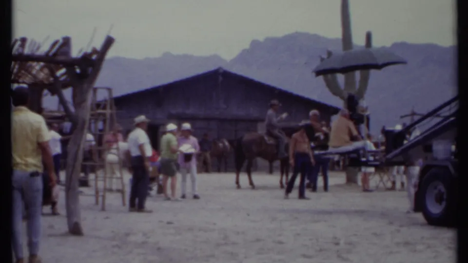 1971 California 8mm still showing people clustered near a horse, ladders, a large umbrella, a vehicle edge, and a rustic set facade.