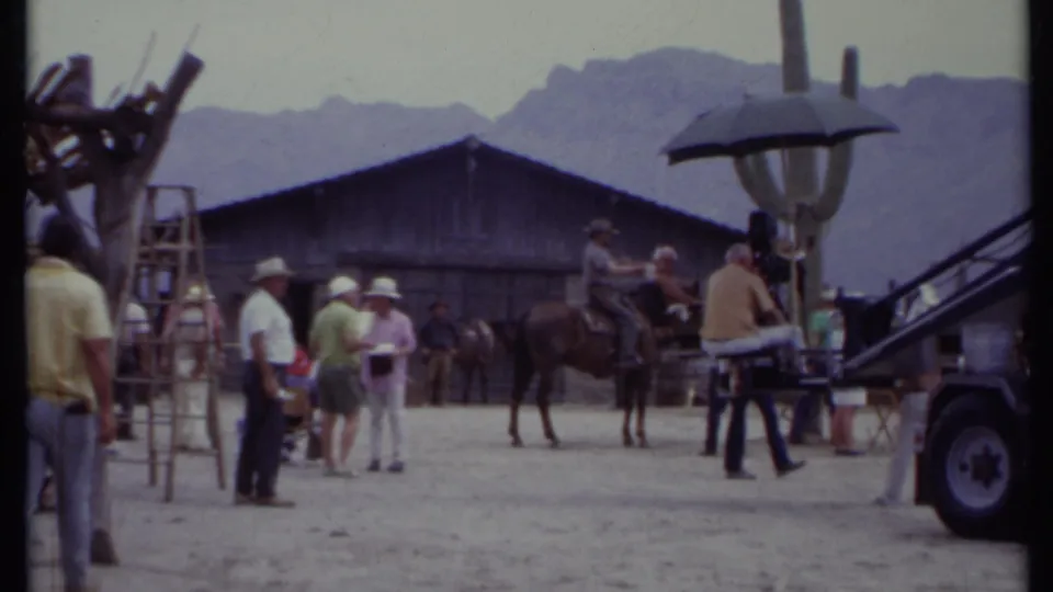 1971 California frame showing a Western-style horseback setup with nearby crew, dolly or camera rig, umbrella, ladders, and mountains.