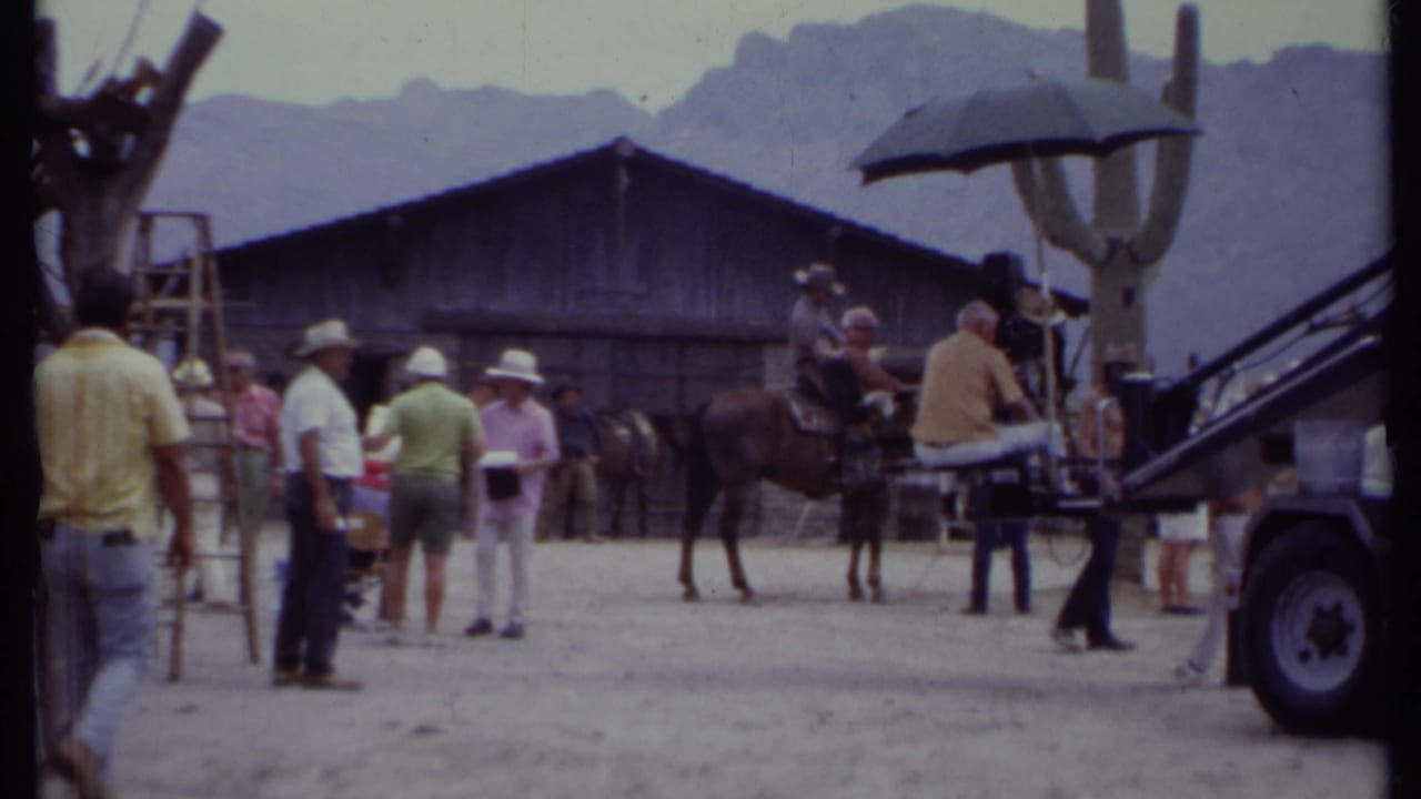 1971 California 8mm frame showing a Western set with crew, a horse, rustic barn, saguaro cactus, sun umbrella, and mountains.