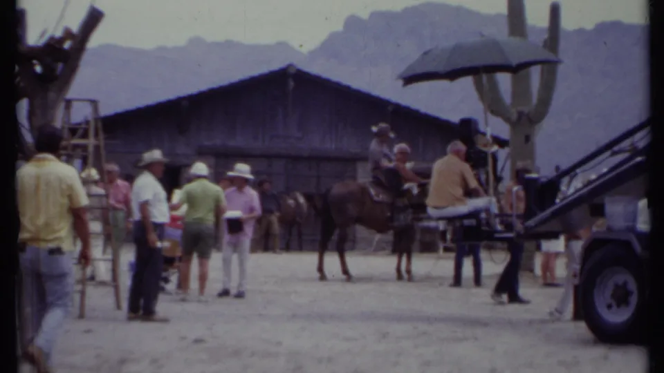 1971 California 8mm frame showing a Western set with crew, a horse, rustic barn, saguaro cactus, sun umbrella, and mountains.