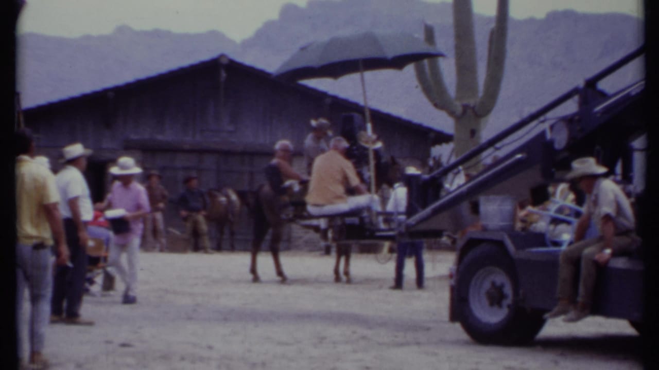 Camera crane vehicle and crew stand near a horse, cactus, umbrella, and wooden set building.