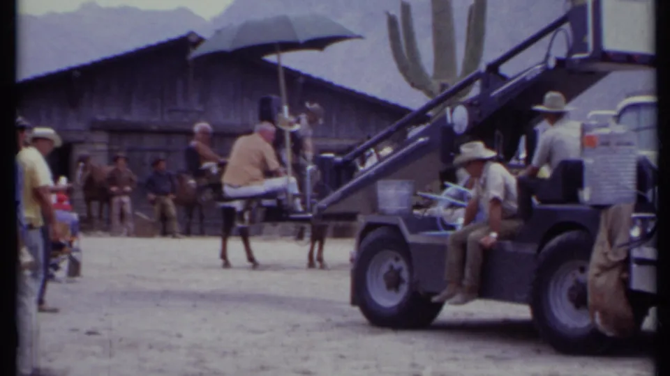 1971 California 8mm frame showing the crane vehicle closer to the Western set, with crew, horse, cactus, umbrella, and mountains behind.