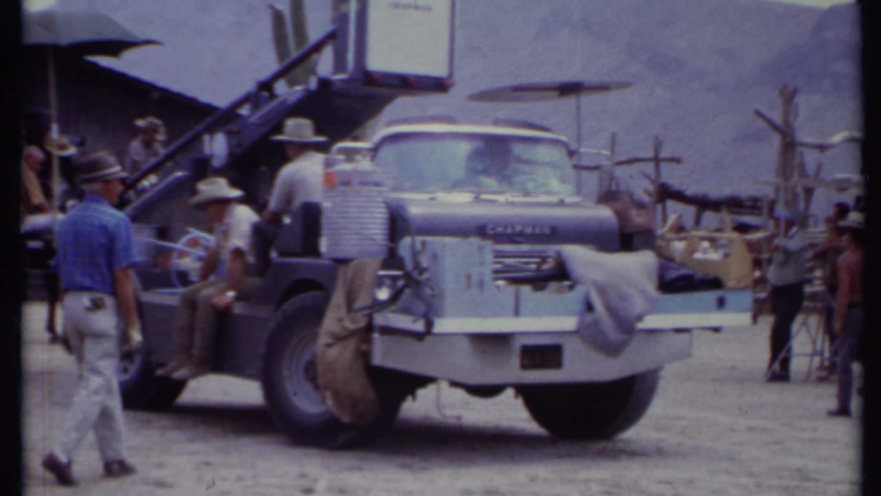 Blue equipment truck at a rustic outdoor set with crew members in hats nearby.