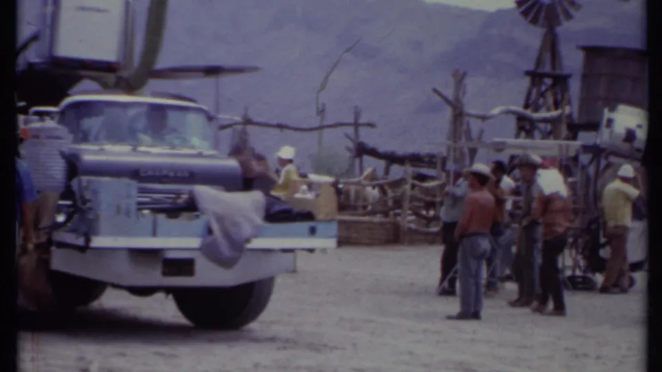 1971 California frame with a loaded truck, people in hats, wooden set rails, windmill, water tank, and mountain backdrop.