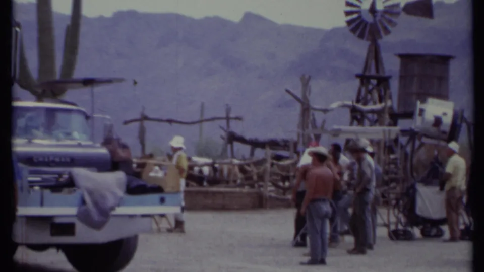 1971 California 8mm frame showing crew beside production equipment, rustic set fencing, a windmill, and hazy mountains.